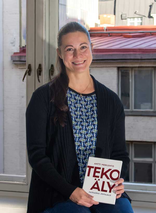 Maria Ojajärvi istuu ikkunalaudalla Tekoäly-kirja kädessään ja kaupungin sisäpihanäkymä taustalla.
