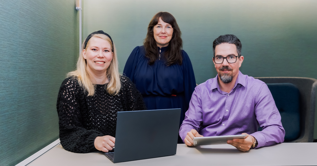 Kolme henkilöä istuu pienen neuvotteluhuoneen pöydän ääressä. Yhdellä on avoin kannettava tietokone, toisella tabletti. Tilanne viittaa yhteiseen työskentelyyn tai palaveriin.