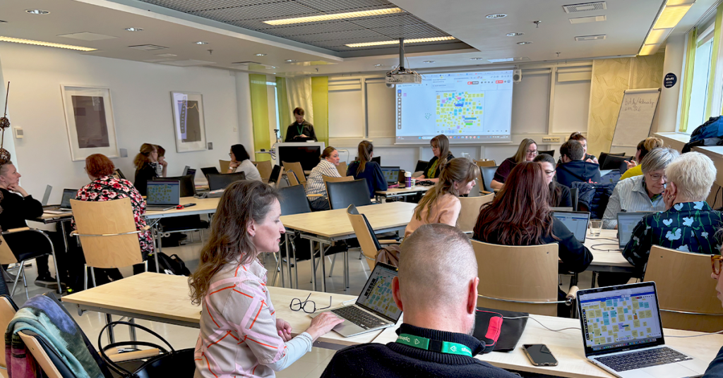 Luokkahuonemaisessa tilassa useita henkilöitä työskentelee pienryhmissä kannettavien tietokoneiden äärellä. Etualalla henkilöitä istuu pöytien ääressä katsomassa läppärien näyttöjä, joilla näkyy värikkäitä muistilappuja muistuttava digitaalinen ideointialusta. Taustalla tilan etuosassa henkilö seisoo puhujakorokkeen takana, ja seinälle projisoidulla valkokankaalla näkyy sama digitaalinen ideointinäkymä. Huoneessa on vaaleat seinät, kattovalaisimet ja ikkunoista tulevaa luonnonvaloa.